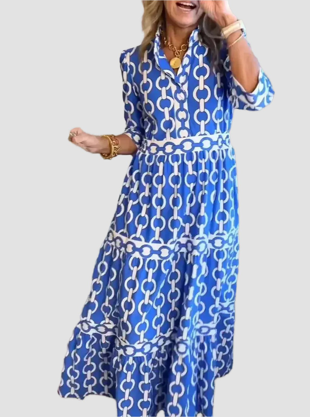 A woman wearing a blue and white patterned dress with a high collar and long sleeves, smiling and gesturing with her hand.